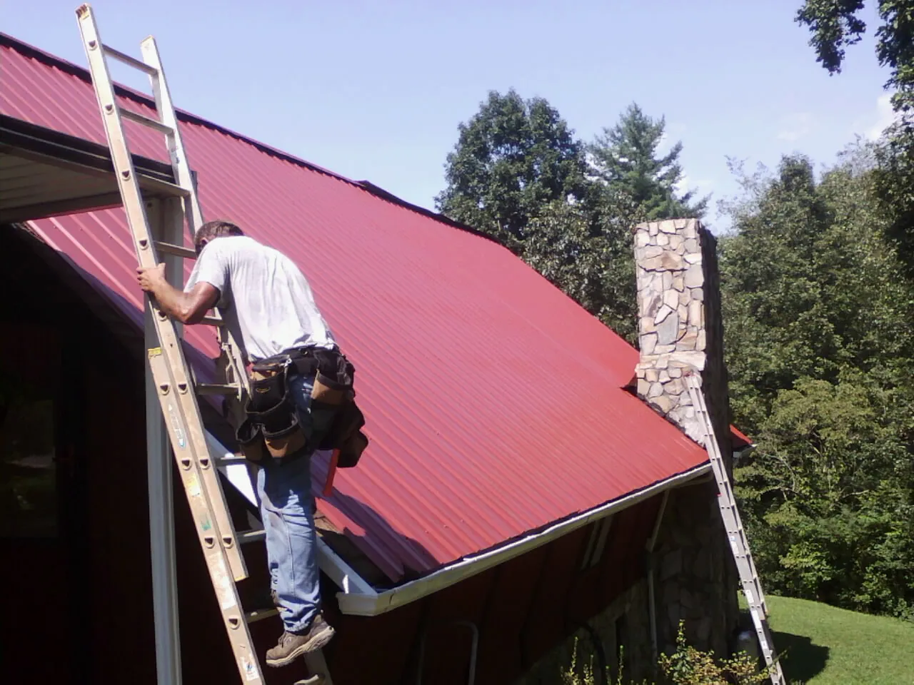 Roofer on a ladder beside a red metal roof and stone chimney — Soffit & Fascia Repair by Roofing Brothers in Paterson